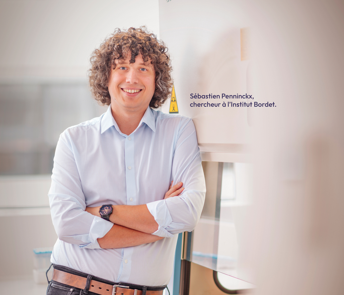 Un homme avec des cheveux bouclés se tient debout dans un laboratoire, avec la mention : "Sébastien Penninckx, chercheur à l'institut Bordet".